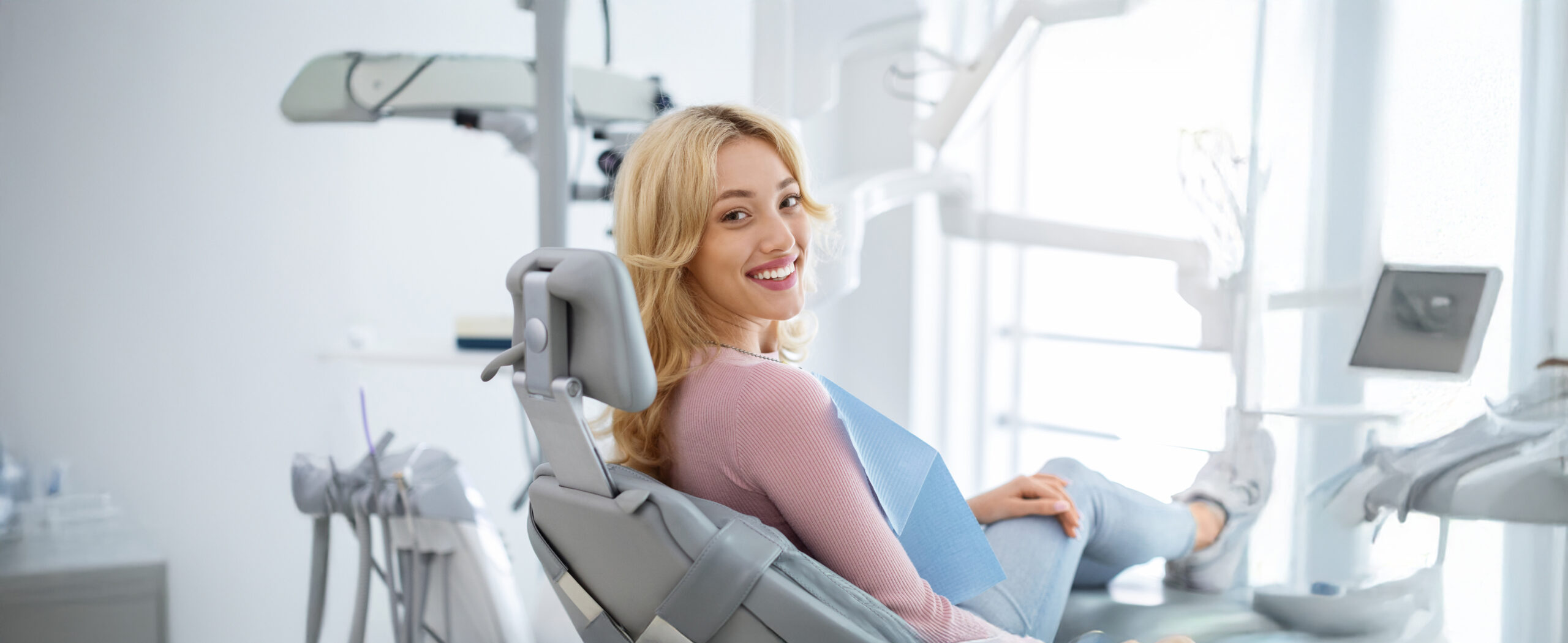 Smiling and relaxed young woman sitting at dental chair in modern clinic, waiting for her dentist. Happy attractive blonde lady attending luxury dental clinic, bacl view, copy space